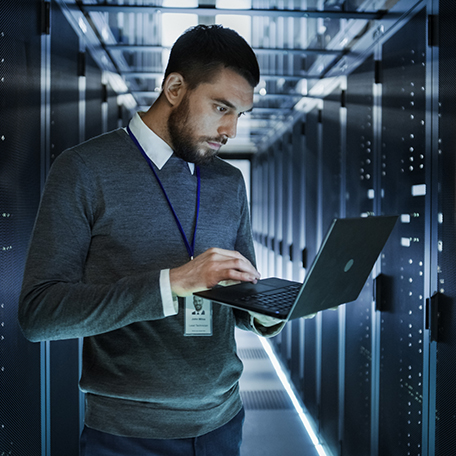 IT Technician Works on a Laptop in Big Data Center full of Rack Servers. He Runs Diagnostics and Maintenance, Sets up System.