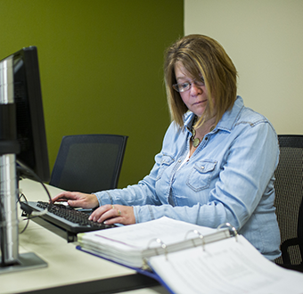 Student working on her desktop computer against a green backdrop, a book lies open on the table