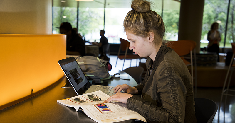 Student sitting at a table working on a laptop with a book open