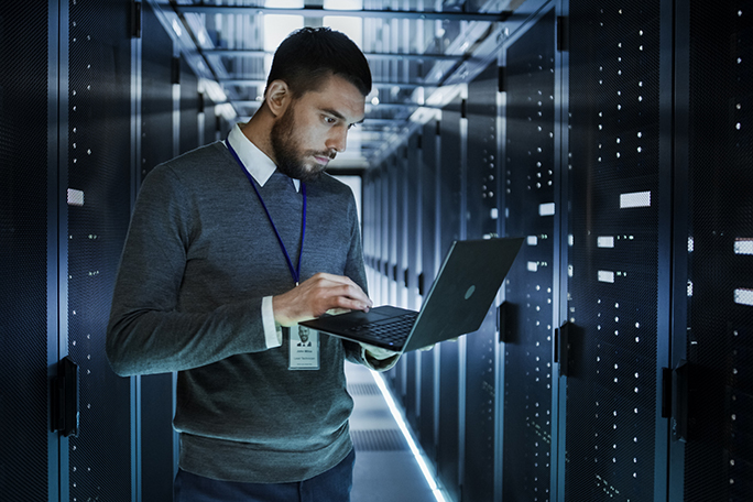 IT Technician Works on a Laptop in Big Data Center full of Rack Servers. He Runs Diagnostics and Maintenance, Sets up System.