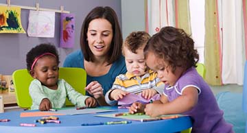 A toddler caregiver looking at the artwork of three toddler-aged kids in a school-like setting
