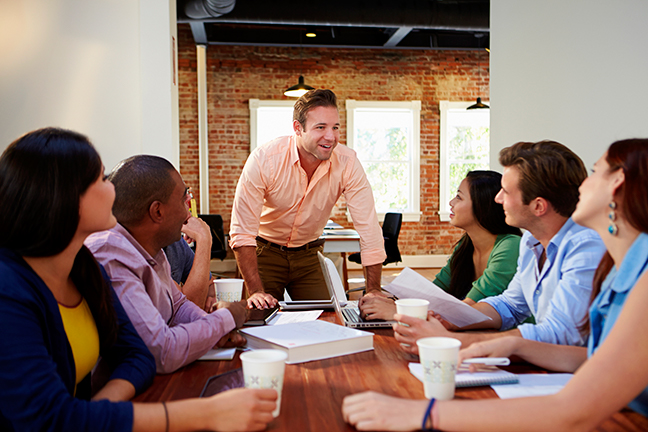 A meeting with a group of professionals and the leader standing at the head of the table looking very excited