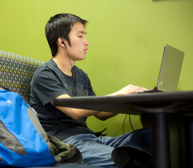 Young, male student working on his computer in a booth on campus with headphones in.