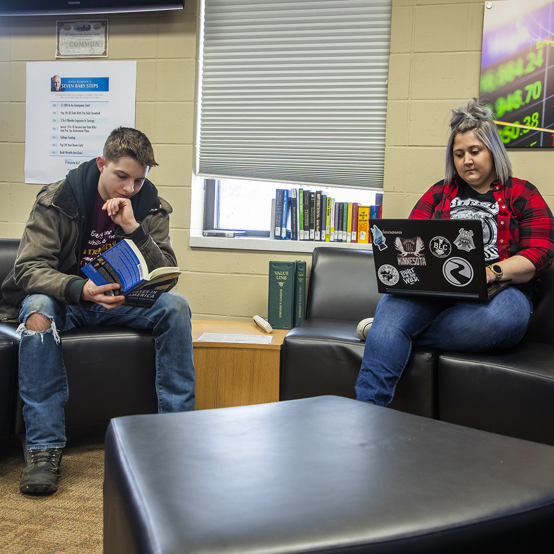 Two students in a classroom. One student is reading and another student is using their laptop