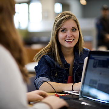 Student working at a laptop and looking up and smiling at a classmate