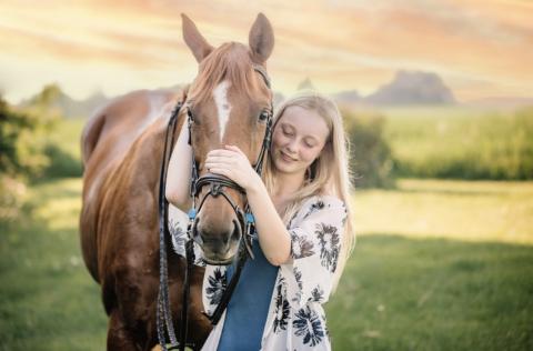 Cassandra Gulich smiles while gently hugging a chestnut horse in a sunny field at sunset.