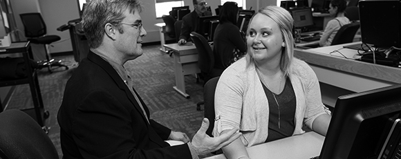 A finance instructor sits with a young female student troubleshooting a problem on her computer in the modern finance classroom.