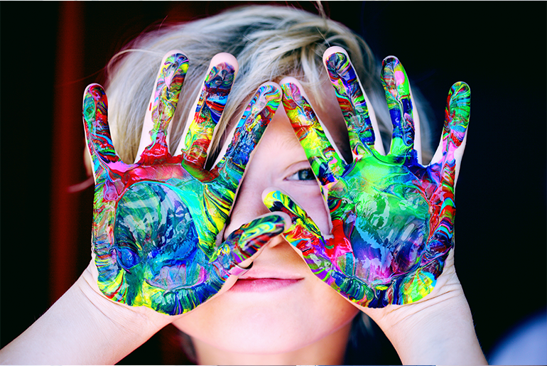 Close up of a five year old boy holding his paint colored hands palms out, in front of his face