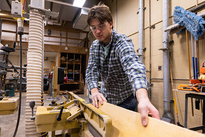 Person guiding a wooden board through a jointer in a woodworking shop.