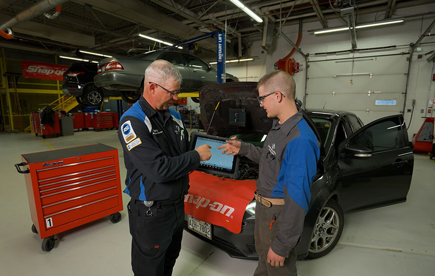 Instructor and student using a diagnostic tool on a car in an automotive workshop.