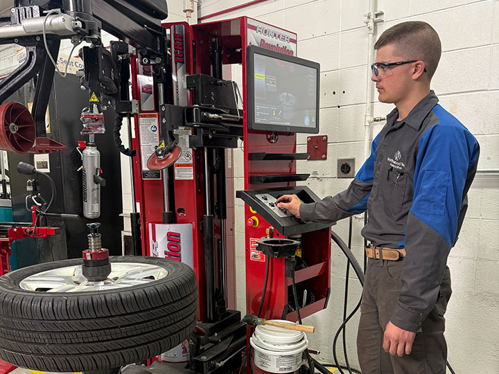 Student operating a tire mounting machine with a wheel attached in an automotive workshop.
