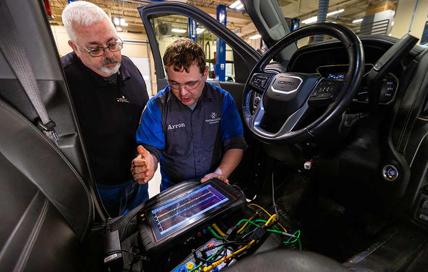 Two individuals using a diagnostic scanner inside a vehicle to check electronic systems in an automotive workshop.