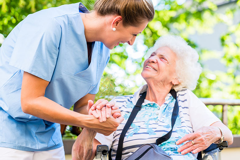 Community Based Residential Facility Caregiver holds hand and smiles with an older female in a wheelchair.