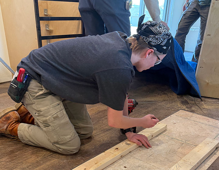 Student kneeling on the floor, securing a wooden board with a drill during a construction project.