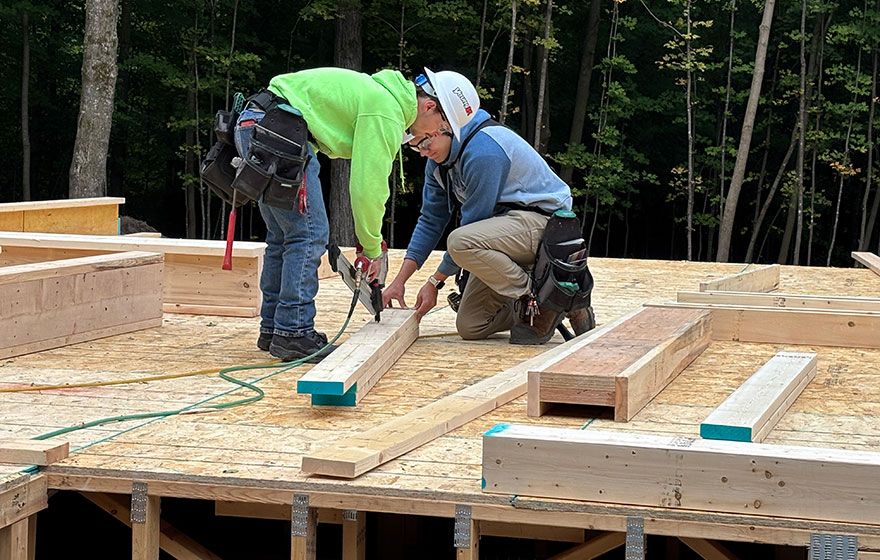 Two individuals working on a wooden structure, measuring and securing boards at a construction site.