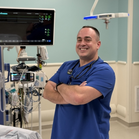 Man wearing blue medical scrubs stands in a hospital room with arms crossed, smiling beside medical monitoring equipment.