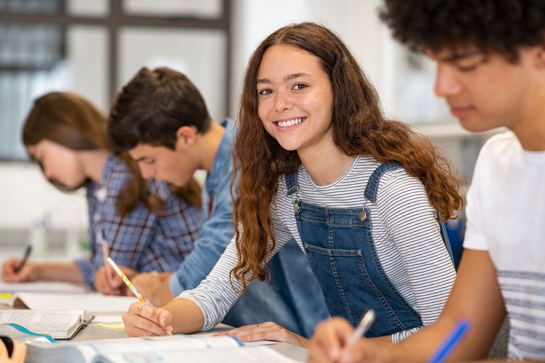 Student holding a pen in a class room with other students, smiling