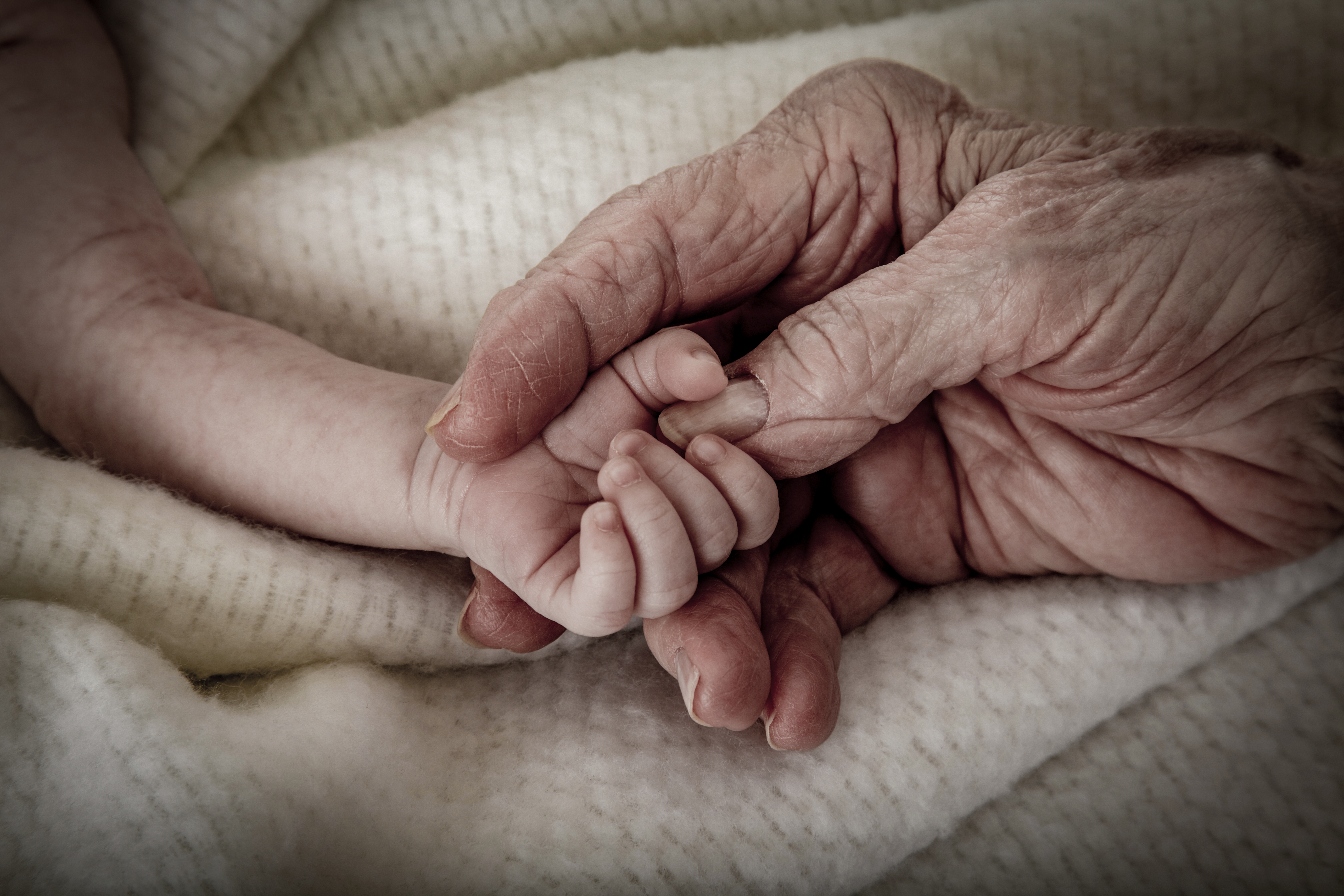 An older person’s hand gently holding a baby’s hand on a soft blanket.