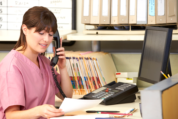 A medical professional talking on the phone at a desk with folders behind her