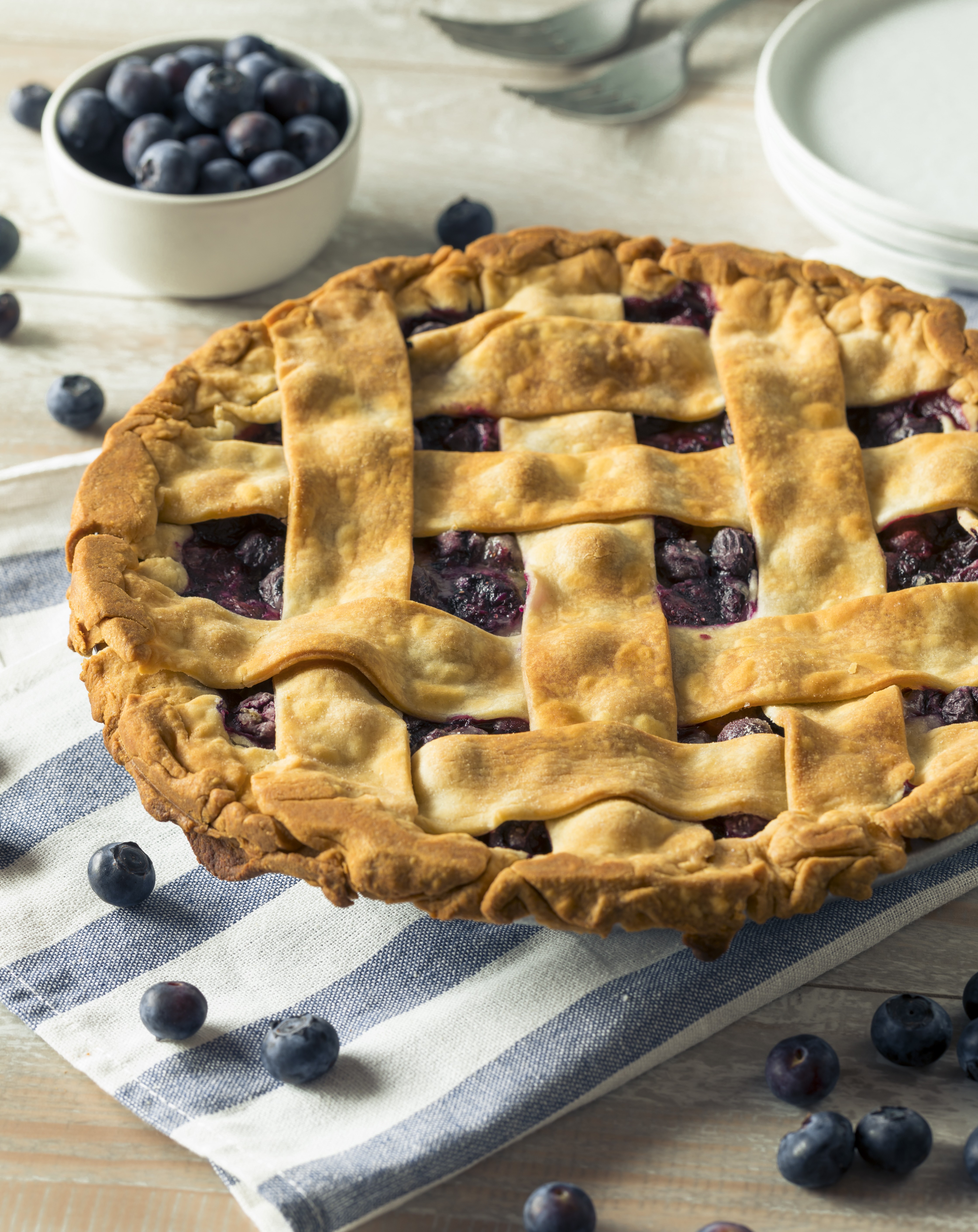 A golden lattice blueberry pie sits on a striped cloth with scattered blueberries, a small bowl, and plates in the background.