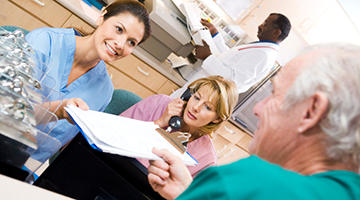 A busy clinic front desk with a receptionist handing paperwork over to a patient