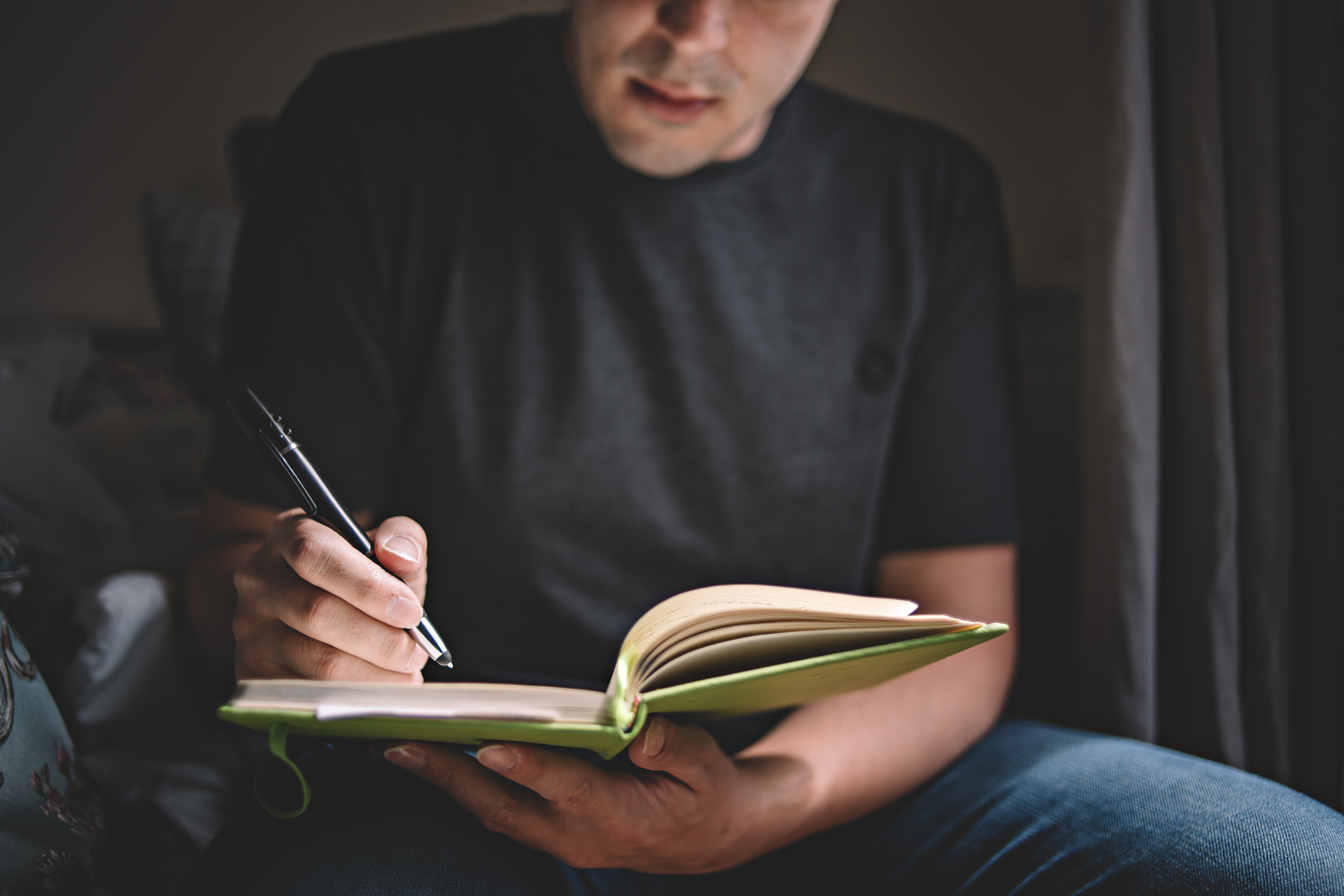 Person seated indoors writes in an open notebook with a pen, holding the journal in their hands.
