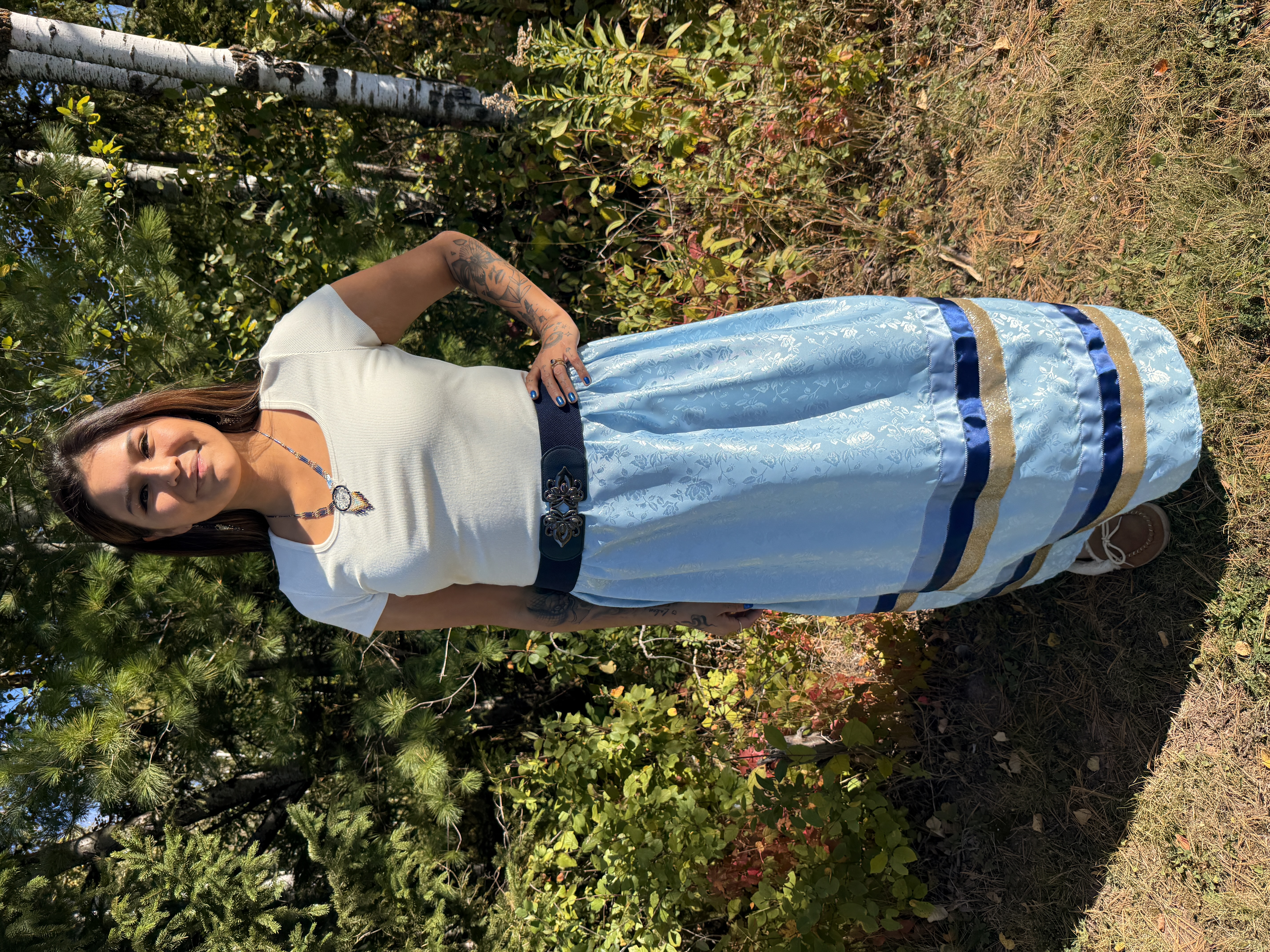 Jami Pierre, a member of the Fort William First Nation, stands outdoors surrounded by trees and fall foliage, wearing a white to