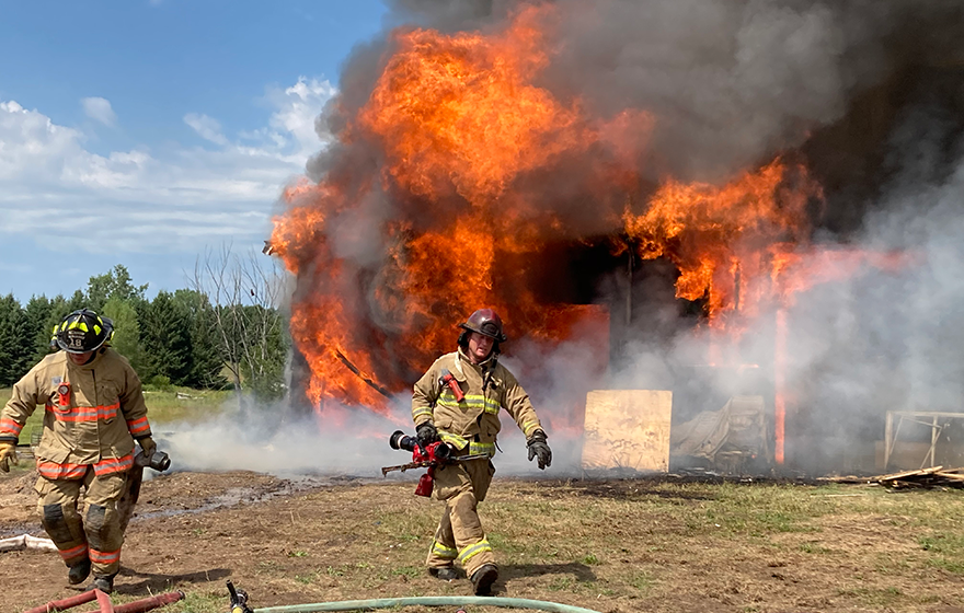 Firemen in full gear practice with house on fire in background