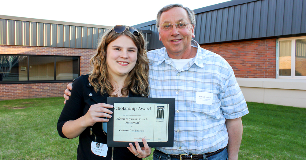 Female student standing outside holding a scholarship plaque. She is standing next to the man who is the scholarship donor.