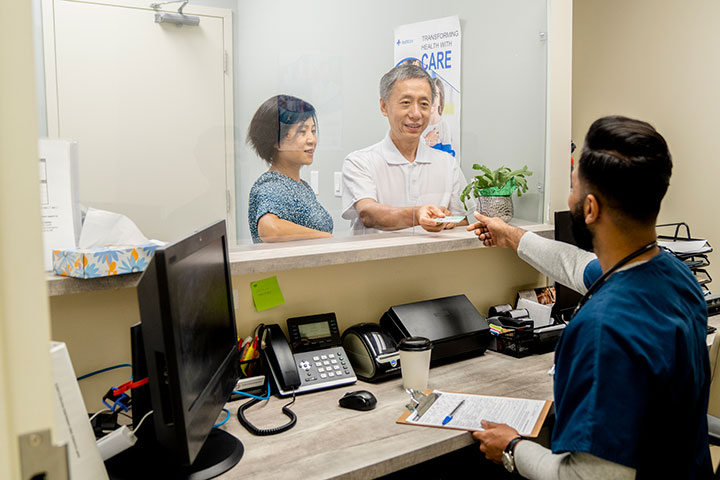 Medical administrative professional handing paperwork to two people at a clinic reception window.