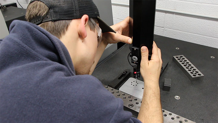 Student using a coordinate measuring machine to inspect a metal part in a workshop setting.
