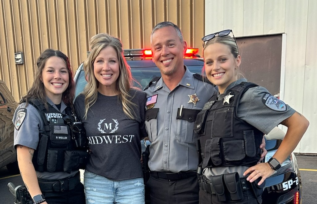 Four adults smile while standing together in front of a sheriff’s patrol vehicle with emergency lights visible.  