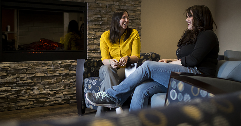 Two female students sitting by fireplace laughing and sitting on furniture 