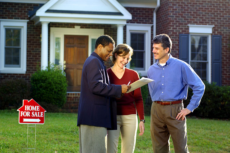 Real Estate agent reviews paperwork with a couple on the front lawn of a home with a for sale sign nearby