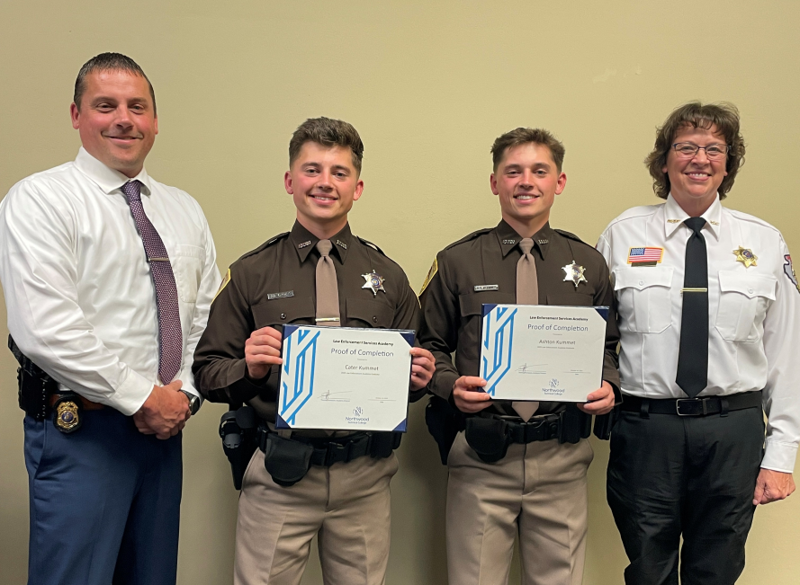 Four adults stand indoors, with two individuals holding certificates and two others standing beside them.