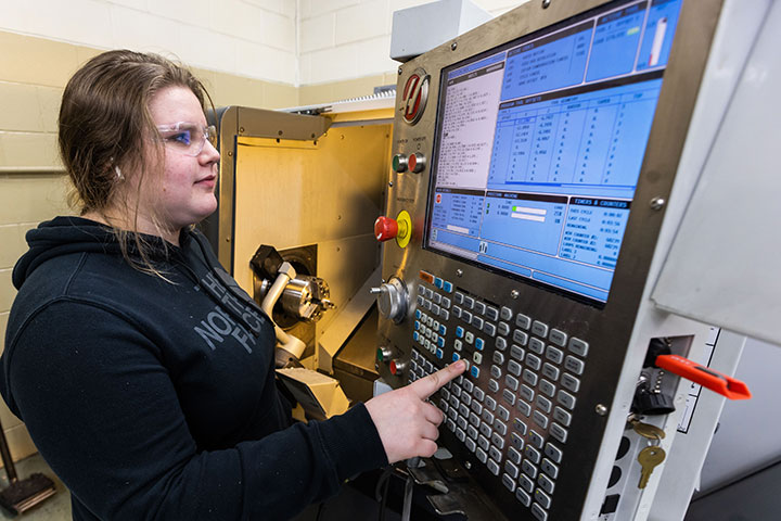 Female student operating a CNC machine, pressing buttons on a control panel with screens displaying technical data.