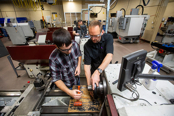 Instructor guiding a student on a lathe machine in a workshop with multiple CNC machines in the background.