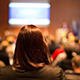 The back of a woman's head while she faces a crowd and a presenter in a dimly lit conference center at an event