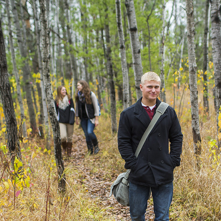 Three students, a male and two females, walking outside on a wooded trail in the fall.