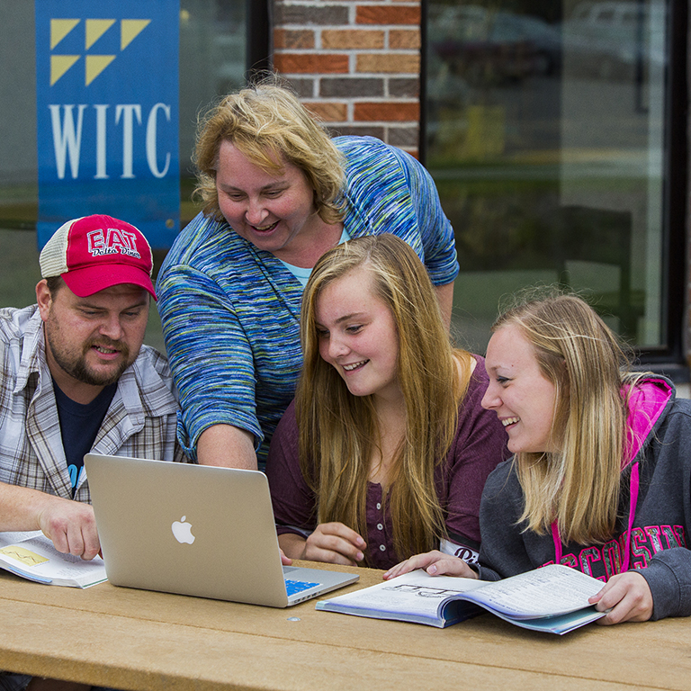 A group of individuals, one male and two females, gathered around a computer and books with an instructor overlooking to assist.