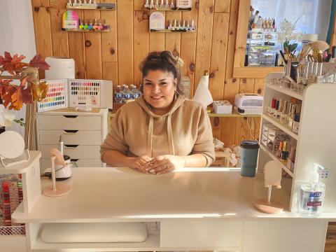 Rachel Aranda smiles while seated at her nail station inside Polished Beauty & Spa with shelves of colorful nail polishes behind