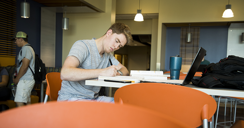 Student works at his computer in the student lounge and write in his notebook