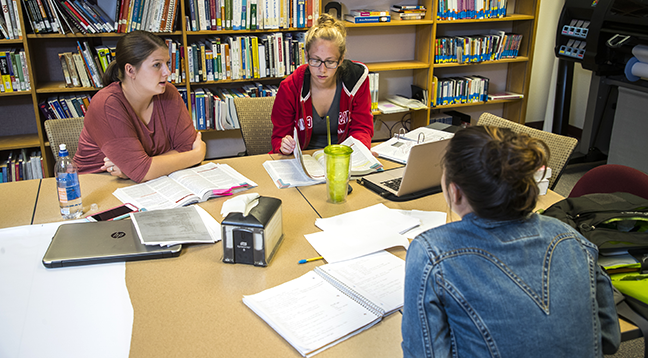 Students studying in a library at a table with papers spread around