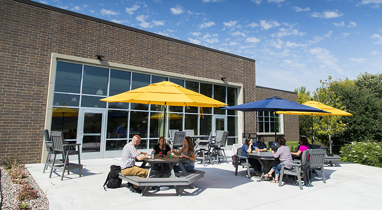 Students sit out on a bright sunny patio at table with blue and gold umbrellas. 